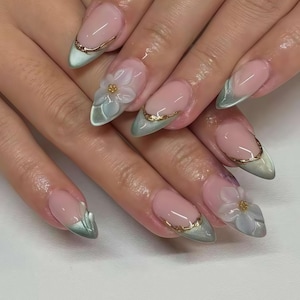 May include: Oval-shaped nail art with a light pink base, accented with a gold trim and a gradient of light green at the tips. Some nails feature a 3D white flower with a gold center, creating a delicate and elegant design.