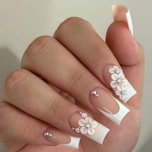 Puede incluir: Una mano que muestra uñas acrílicas largas y cuadradas. Las uñas presentan un diseño de manicura francesa con puntas blancas y una base nude. Cada uña está adornada con adornos florales blancos en 3D y pequeños pedrería brillantes.