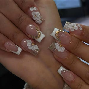 Puede incluir: Una manicura con uñas acrílicas largas y cuadradas. Las uñas presentan un diseño de punta francesa con puntas blancas y una base nude. Algunas uñas están adornadas con flores blancas en 3D, pedrería dorada y purpurina.
