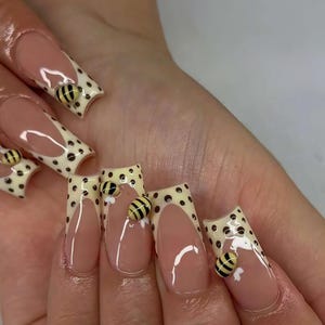 Puede incluir: Primer plano de una mano con uñas acrílicas largas y cuadradas. Las uñas presentan una base beige claro con una punta blanca adornada con lunares negros. Cada uña tiene un dije de abeja en 3D.
