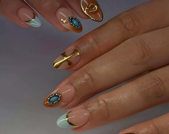 May include: Close-up of manicured nails with a light pink base. Some nails feature gold accents, including a cross, a crescent, and a border. Other nails have a light blue tip with a gold border, and a teal-coloured gem.