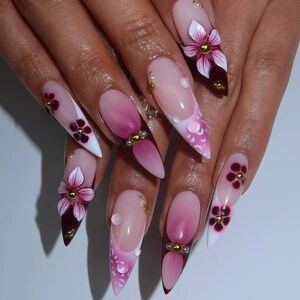 May include: A set of long, pointed acrylic nails with a gradient pink and white design. Each nail features 3D floral embellishments in shades of pink and burgundy, accented with gold beads and clear droplets.