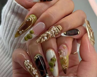 Puede incluir: Una mano que muestra uñas acrílicas largas en forma de almendra. Las uñas presentan una variedad de diseños, incluyendo detalles de alambre dorado, perlas y un patrón de carey. Los colores incluyen marrón, blanco y verde oliva.
