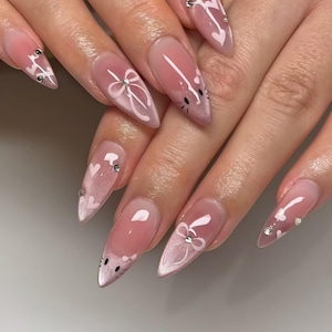 May include: Long, almond-shaped nails with a pink gradient and white designs. The designs include bows, hearts, and cat faces. Some nails have small, silver rhinestones. The nails are well-manicured and polished.