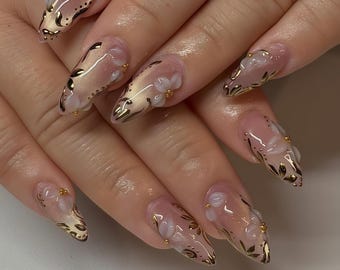 Könnte beinhalten: Ein Set langer, mandelförmiger Kunstnägel. Jeder Nagel hat eine transparente Basis mit einem Farbverlauf aus Rosa und Gold. Zarte weiße Blumenakzente und Goldblattdesigns zieren die Nägel, kleine goldene Perlen fügen Details hinzu.