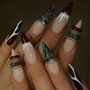May include: A hand with long, pointed nails featuring various designs. Some nails have a nude base with white tips, while others showcase a tortoiseshell pattern. Several nails are adorned with stacked rings in gold, green, and brown tones, some with a small gem.