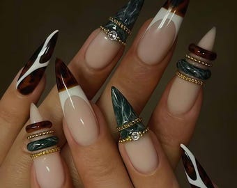 Puede incluir: Una mano con uñas largas y puntiagudas con varios diseños. Algunas uñas tienen una base nude con puntas blancas, mientras que otras muestran un patrón de carey. Varias uñas están adornadas con anillos apilados en tonos dorados, verdes y marrones, algunos con una pequeña gema.