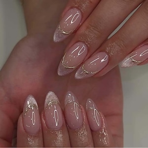 May include: A close-up of a hand with long, oval-shaped nails. The nails are painted with a light pink base and feature a silver glitter tip with a thin gold line. The nails are well-manicured and glossy.