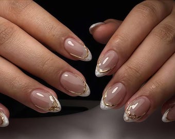 Könnte beinhalten: Nahaufnahme von manikürten Nägeln mit nackter Basis, weißen Spitzen und goldenen Akzenten. Die Nägel haben eine ovale Form mit einer dünnen goldenen Kontur und einem kleinen goldenen Schleifendesign in der Nähe der Nagelhaut. Der Gesamteindruck ist elegant und raffiniert.