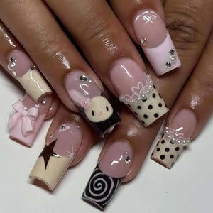 Puede incluir: Un conjunto de uñas artificiales largas y decoradas. Los diseños incluyen un conejito, lunares, lazos y patrones de estrellas. Los colores incluyen rosa, negro, blanco y beige. Algunas uñas tienen pedrería y perlas.