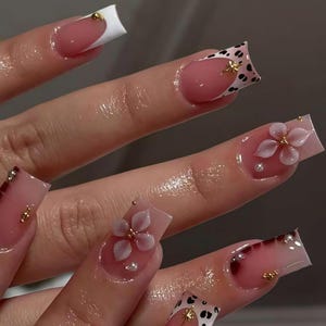 Puede incluir: Un conjunto de uñas acrílicas largas y cuadradas. Las uñas presentan una variedad de diseños, que incluyen una punta francesa blanca, una base rosa con un diseño floral y un patrón de lunares en blanco y negro. También hay detalles dorados y gemas.