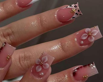Può includere: Un set di unghie acriliche lunghe e quadrate. Le unghie presentano una varietà di design, tra cui una french manicure bianca, una base rosa con un motivo floreale e un motivo maculato bianco e nero. Sono presenti anche dettagli dorati e gemme.