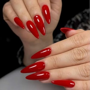 Könnte beinhalten: Nahaufnahme von Händen mit langen, glänzenden, roten Stiletto-Nägeln. Die Nägel haben eine leuchtende, einfarbige Farbe mit einem Hochglanz-Finish. Die Nägel sind gut manikürt und geformt und reflektieren das Licht.