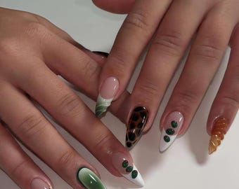 Puede incluir: Un conjunto de uñas acrílicas largas en forma de almendra. Los diseños incluyen un remolino verde y blanco, un estampado de leopardo, puntos verdes en una punta blanca, un acento metálico dorado y un diseño ámbar transparente texturizado.