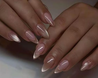 Könnte beinhalten: Nahaufnahme von manikürten Nägeln. Die mandelförmigen Nägel haben eine hellrosa Basis und eine schimmernde, irisierende weiße Spitze. Eine dünne, metallisch goldene Linie umrandet das Nagelbett.
