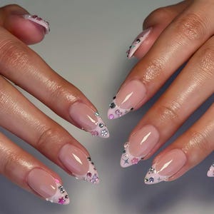 May include: Close-up of a set of long, almond-shaped nails. The nails have a nude base with a French tip design. The tips are decorated with small, sparkling rhinestones and pink and silver accents.