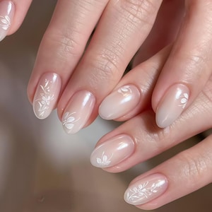 May include: Close-up of a hand with oval-shaped nails. The nails are painted with a gradient of light pink to a nude color. Each nail features a delicate white floral design, adding a touch of elegance to the manicure.