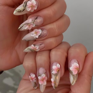 Puede incluir: Un conjunto de uñas artificiales largas en forma de almendra. Cada uña presenta una base rosa claro con un diseño floral en 3D. Las flores son rosas y blancas con detalles dorados, creando un aspecto elegante y artístico.