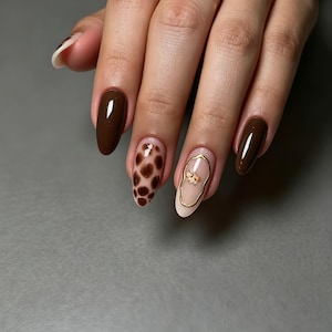 May include: Almond-shaped fingernails featuring a mix of solid dark brown polish, a brown spotted animal print pattern on a nude base, and a nude nail with a gold metallic outline surrounding a small, delicate gold flower embellishment.