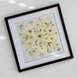 May include: A framed floral arrangement featuring a variety of dried flowers in shades of white and cream. The flowers are arranged in a circular pattern within a white mat, encased in a dark brown wooden frame. The artwork is displayed on a white surface.