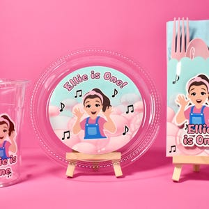 May include: A clear plastic cup with a pink and white striped straw, a plate, and a utensil holder. Each item features a cartoon girl and the text "Ellie is One!" on a pink background. Musical notes are also present.