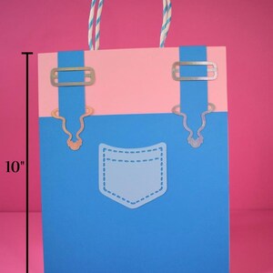 May include: A large gift bag with a blue and pink design, resembling denim overalls. The bag is 10 inches tall and 8 inches wide, with a white pocket outline and silver-colored metal accents. The handle is a twisted white and blue cord.
