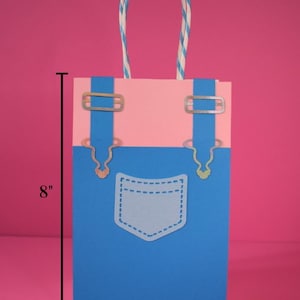 May include: A medium-sized gift bag, 8 inches tall and 5 inches wide, designed to resemble denim overalls. The bag features a blue lower section with a pocket, a pink upper section, and silver-colored hardware accents. The handle is striped.