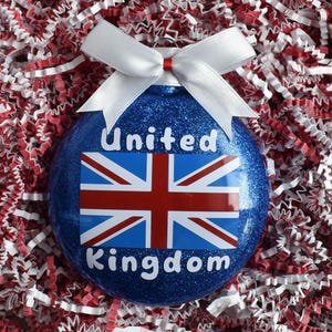 May include: A blue glitter Christmas ornament with the Union Jack flag and the words "United Kingdom" in white. The ornament has a white bow at the top and is set against a background of red and white shredded paper.