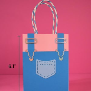 May include: A small, paper gift bag with a denim overalls design. The bag is blue and pink, with a white pocket and silver-colored metal accents. It measures 6.1 inches tall and 4.5 inches wide. The handles are white and blue striped.