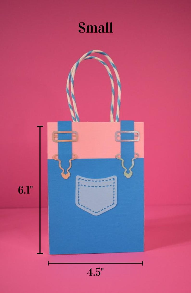May include: A small, paper gift bag with a denim overalls design. The bag is blue and pink, with a white pocket and silver-colored metal accents. It measures 6.1 inches tall and 4.5 inches wide. The handles are white and blue striped.