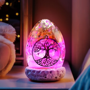 Pode incluir: Uma luz noturna em forma de ovo com um design de árvore da vida. A lâmpada tem um interior rosa e dourado com glitter e assenta numa base texturizada em branco sujo. A lâmpada está numa mesa de cabeceira branca.