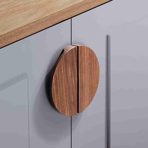 Puede incluir: Un tirador de armario circular hecho de madera marrón con un patrón de veta visible, dividido en dos mitades semicirculares. El tirador está montado en una puerta de armario gris claro con acabado liso, situada bajo una encimera de madera clara.