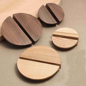 Puede incluir: Cuatro soportes circulares de madera, cada uno con una ranura horizontal recta en el centro. Dos soportes están hechos de madera marrón oscura con vetas visibles, y dos están hechos de madera color canela claro con un patrón de vetas naturales.