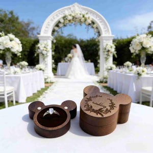 Peut inclure: Deux boîtes à bagues en bois en forme de tête de souris, l'une ouverte avec une bague en diamant, l'autre fermée avec le mot "wedding" gravé sur le couvercle. Les boîtes sont sur une surface blanche avec une cérémonie de mariage en arrière-plan.