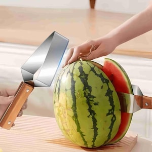 Puede incluir: Un cortador de sandía de acero inoxidable con mango de madera corta una sandía a rayas verdes y negras. Se ve una rodaja roja de sandía. El cortador es sostenido por una mano.