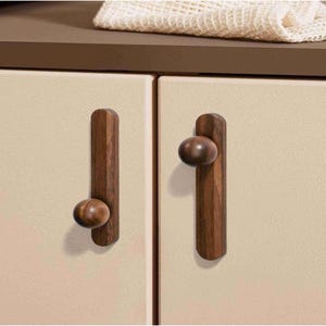 May include: Two cabinet doors with wooden pulls. Each pull has a dark brown, elongated base with a rounded knob. The cabinet doors are a light beige colour. A beige cloth is visible on the countertop above.