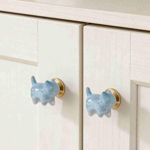 May include: Two light blue cat-shaped cabinet knobs with brass-colored bases are attached to a white cabinet. The knobs have a marbled appearance with shades of blue and white. The cabinet doors are made of wood.