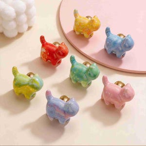 May include: A collection of colorful ceramic cat-shaped cabinet knobs. The knobs are in various colors, including red, yellow, blue, green, and pink. Each cat has a gold-colored base for mounting. The knobs are designed for furniture decoration.