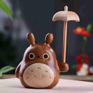 May include: A wooden figurine of a cartoon character with a round body, large ears, and a light-colored belly. The figurine is holding a small wooden umbrella. The character has large, round eyes and a friendly expression. The wood has a warm, brown tone.