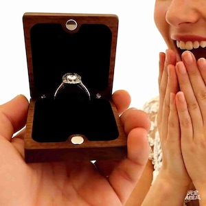 May include: A silver engagement ring with a halo of smaller stones is displayed in an open, wooden ring box with a black velvet interior. The box is held in a person's hand. The ring is the focal point.