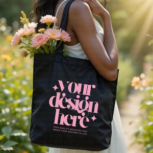 May include: A black tote bag with pink text reading "YOUR design here", filled with pink flowers. The bag is held by a person in a white dress, with a blurred background of greenery and sunlight. The bag also has the text "HIGH QUALITY MOCKUPS THAT BRING YOUR DESIGNS TO LIFE".