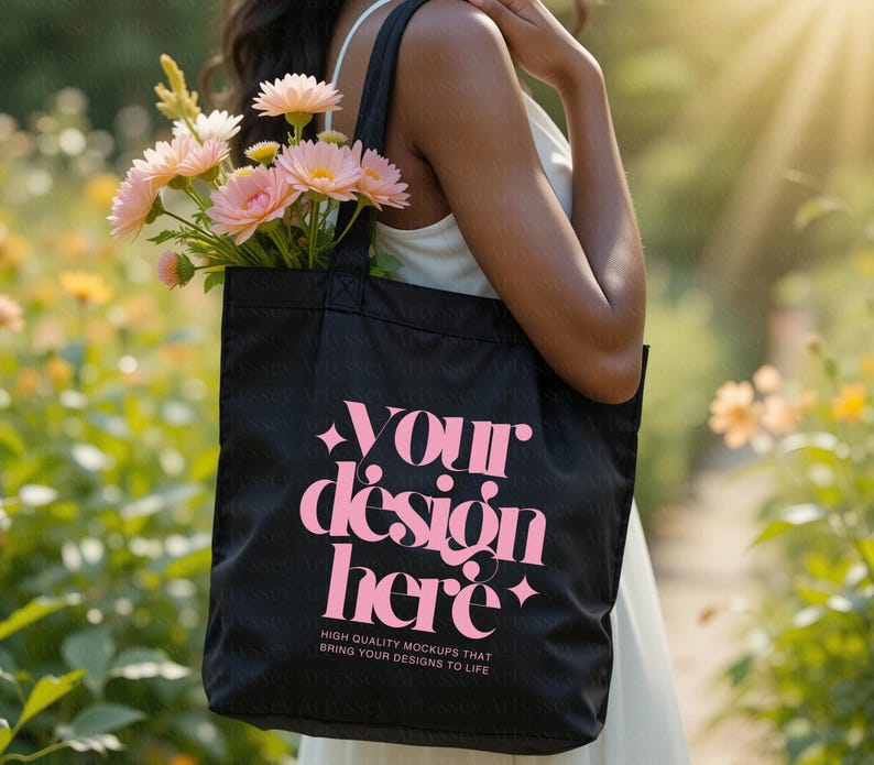 May include: A black tote bag with the text "YOUR design here" in pink, filled with pink flowers. The bag is held by a person in a white dress, with a blurred background of greenery and sunlight. The bag also has the text "HIGH QUALITY MOCKUPS THAT BRING YOUR DESIGNS TO LIFE".