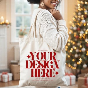 May include: Cream-colored tote bag with the text "YOUR DESIGN HERE" in red, held by a person. The bag has long straps and a rectangular shape. The background includes a Christmas tree and a fireplace.