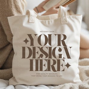 May include: Cream-colored tote bag with the text "YOUR DESIGN HERE" in a brown, bold font. The bag has two handles and is made of a sturdy fabric. The words "HIGH QUALITY MOCKUPS THAT BRING YOUR DESIGNS TO LIFE" are printed at the bottom.
