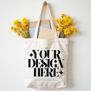 Könnte beinhalten: Cremefarbene Tragetasche mit dem Text "YOUR DESIGN HERE" in fetten schwarzen Buchstaben. Die Tasche ist mit Notizbüchern und gelben Blumen gefüllt. Die Tasche hängt an einem Holzhaken vor weißem Hintergrund.