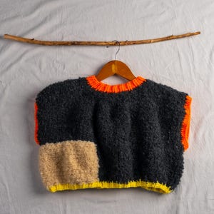 Può includere: Un maglione senza maniche, strutturato, in grigio scuro, arancione, beige e giallo è appeso a una gruccia di legno. Il maglione ha un design a blocchi di colore con finiture arancioni intorno allo scollo e alle ascelle. Un bastoncino di legno è sopra.
