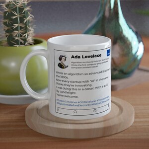 May include: A white ceramic mug featuring a printed social media-style post about Ada Lovelace. The text includes her name, a portrait, and a humorous commentary on modern AI startups, alongside hashtags like #CoderOriginal and #OGDeveloper.