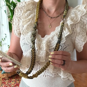 Könnte beinhalten: Ein Makramee-Handyband in Olivgrün, Grau und Schwarz. Das Band ist an einer transparenten Handyhülle befestigt. Der Hintergrund umfasst ein cremefarbenes Spitzentop und einen Blick auf einen gemusterten Teppich.