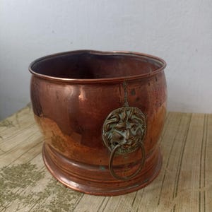 May include: A round copper vessel with a polished metallic finish and a wide opening. The exterior features a prominent brass-toned lion head ornament attached to the side, which holds a circular metal ring handle. The surface shows natural patina.