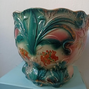 May include: A ceramic planter featuring a scalloped rim and embossed leaf motifs in shades of teal and green. The body displays a cream-coloured background with painted orange floral clusters and subtle pink accents, finished with a glossy glaze.